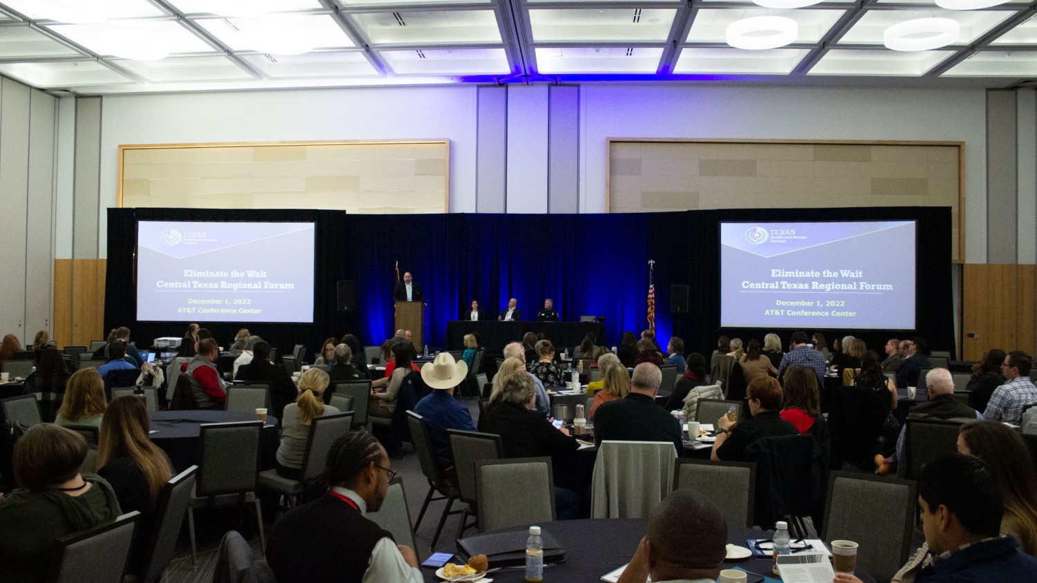 The header image depicts a Texas Health and Human Services conference taking place in a large room. Participants are seated throughout the room, engaged in the event. Two projector screens are positioned on each side of the room, prominently displaying a slide that reads, "Eliminate the Wait Central Texas Regional Forum,” and the date and location of the event. In the center of the room, an individual stands at a podium, delivering a speech to the audience, and three others are seated on stage assuming a speaker role throughout the conference. This photo captures the dynamic of valuable insights and discussions related to eliminating wait times for beds in State hospitals.