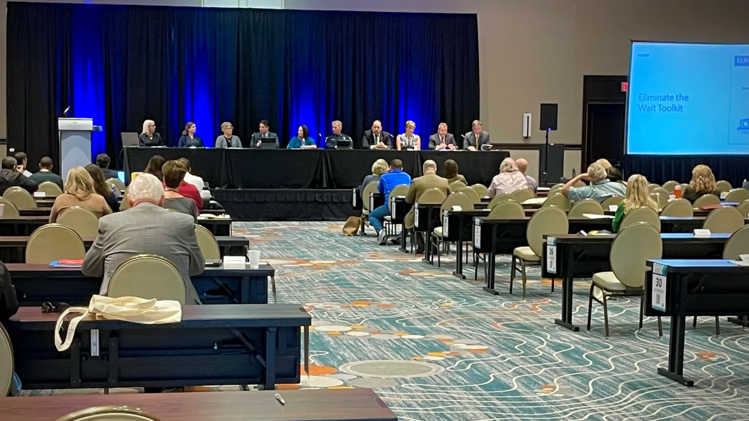 The image captures a conference setting where people have gathered in a room. In the front of the room, a row of individuals is seated facing the crowd, assuming the role of presenters or speakers. The audience is seated in rows of chairs and tables, listening and participating in the event. A large projector screen displays the words "Eliminate the Wait Toolkit." The toolkit aims to address the growing number of individuals within the justice system who require mental health assistance.