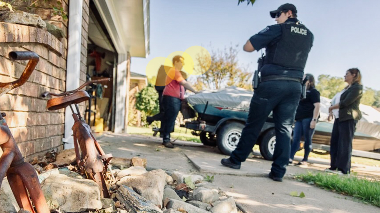 This photo shows four individuals at a private residence. One of these individuals is a police officer, and two others are dressed professionally and holding clipboards. The focus of the image is on the fourth individual, who is possibly the resident of the home. This individual has their hands on the hood of a car that is parked in the driveway. The four people in the image are talking, and the plain clothes people are part of a Community Response Team to provide assistance to the resident of the home.