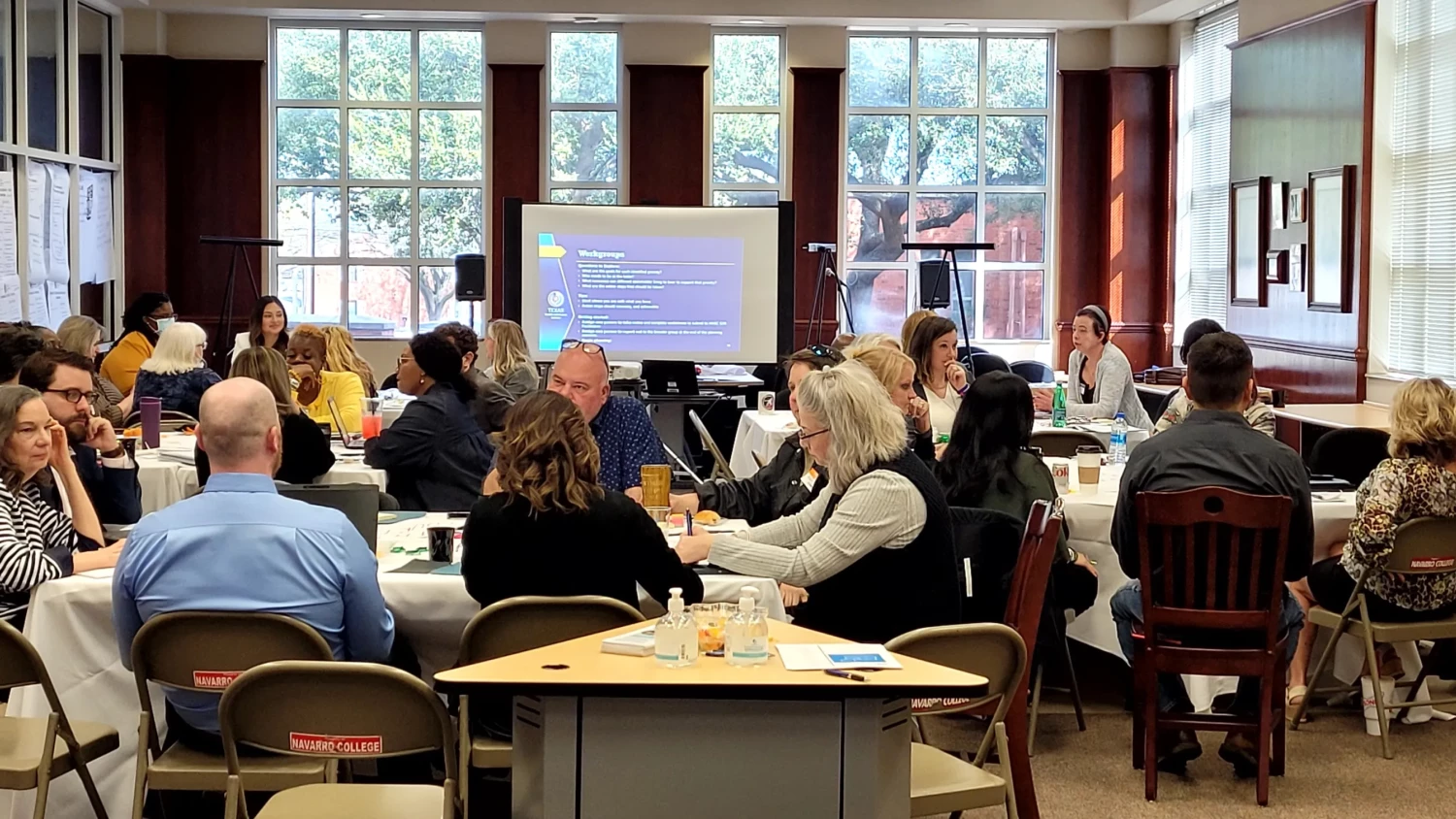 The photo depicts a small room arranged with several round tables where people are seated and engaged in discussions related to the content projected onto a screen at the front of the room. The image was taken at the Navarro College Library during a meeting organized by the North Texas Behavioral Health Authority. The purpose of the meeting is to identify and discuss gaps in the community regarding behavioral health. The participants can be seen actively participating in the conversation, sharing their perspectives, and working together to address the identified gaps.