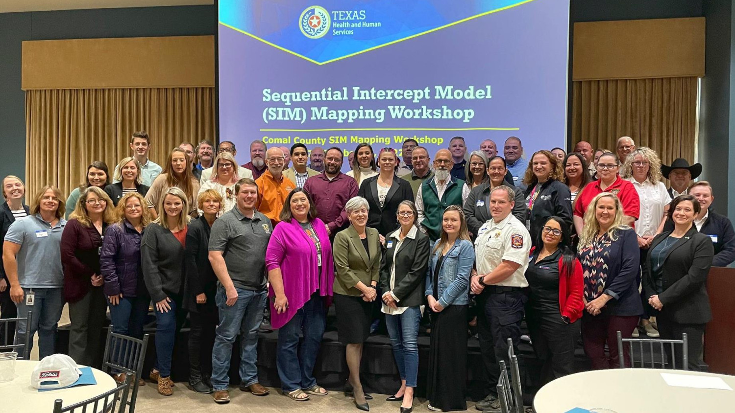 The photo captures a large group of approximately 50 individuals standing together in a unified formation. They are positioned in front of a projector screen displaying the words "Sequential Intercept Model (SIM) Mapping Workshop." The gathering signifies a collective effort to understand and apply the Sequential Intercept Model (SIM) through mapping strategies. The image conveys a sense of collaboration, knowledge sharing, and active participation in the workshop.