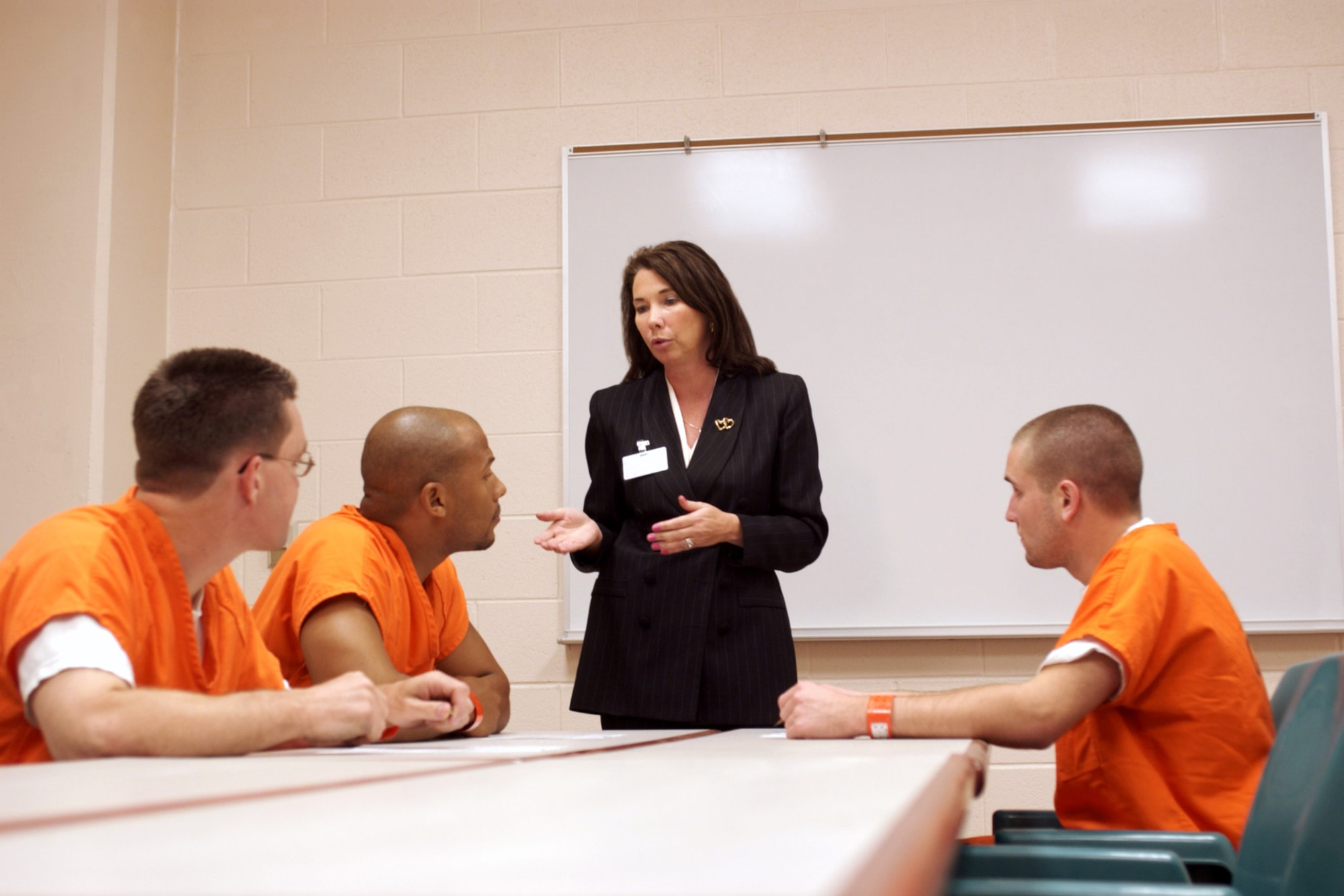 This photo shows four individuals sitting in a room in a correctional facility. An individual dressed in professional clothes is wearing a name tag, standing up and talking with three individuals sitting down at a table wearing orange uniforms. The professional is a social worker) and is counseling the individuals sitting down.
