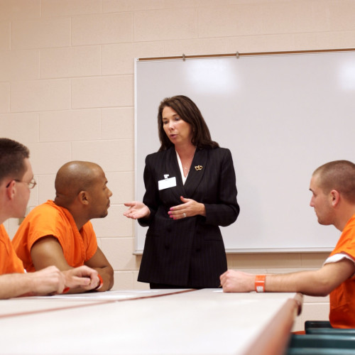 This photo shows four individuals sitting in a room in a correctional facility. An individual dressed in professional clothes is wearing a name tag, standing up and talking with three individuals sitting down at a table wearing orange uniforms. The professional is a social worker) and is counseling the individuals sitting down.