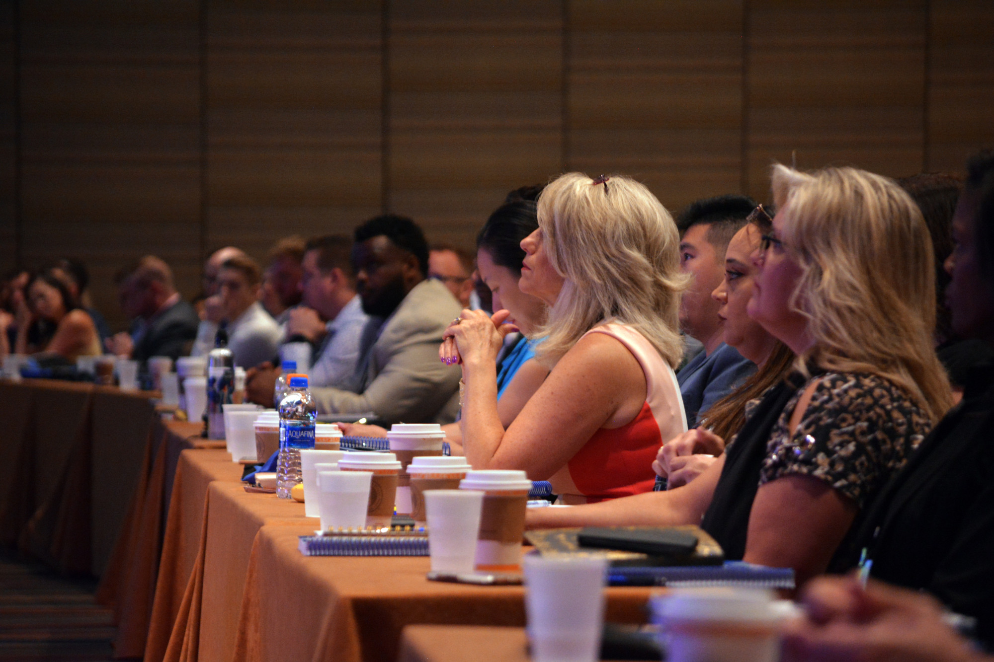 This is a photograph of an audience sitting on chairs at tables with cups of beverages like coffee and water. They appear to be watching and listening to something in front of them, paying attention to it. There is one blonde woman who is the focus of the photo, with others blurred in the distance. It appears they are at a conference, and are attending a presentation.