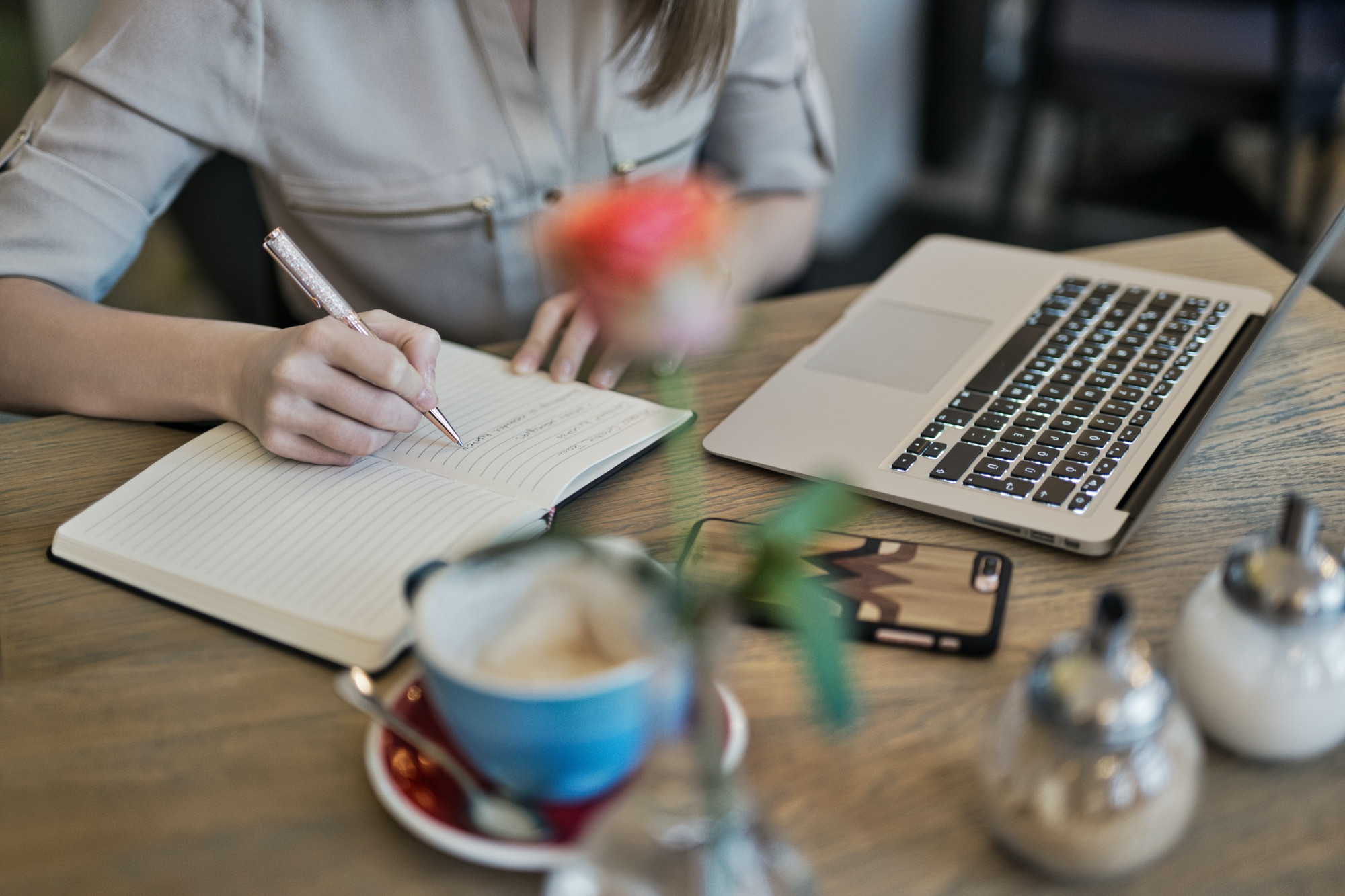 The photo is correlated with the Technical Assistance section of the website and features an individual seated at a table, actively engaged in their work. They are captured writing in a notebook while an open laptop sits nearby. On the table, a coffee mug and a blurred flower add subtle elements to the scene.
