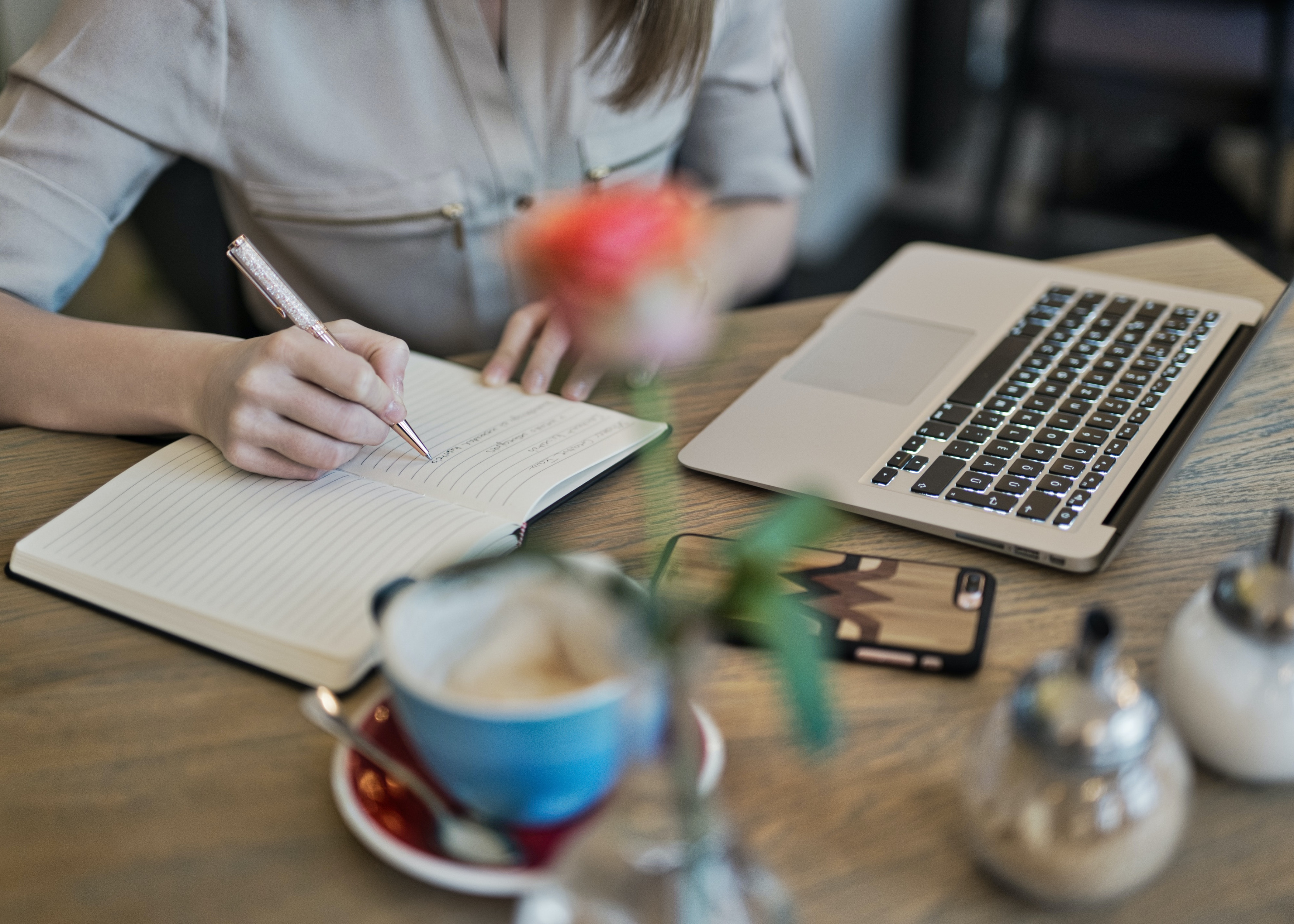 The photo is correlated with the Technical Assistance section of the website and features an individual seated at a table, actively engaged in their work. They are captured writing in a notebook while an open laptop sits nearby. On the table, a coffee mug and a blurred flower add subtle elements to the scene.