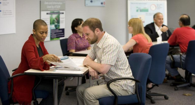 The photo showcases a collaborative learning environment with a room filled with people actively participating. The primary focus is on two individuals situated at the front, who are engrossed in a conversation while examining documents together. The image captures their engaged interaction, highlighting the importance of active communication and collaboration within a learning setting. The presence of other participants in the background suggests a vibrant and inclusive learning atmosphere where shared knowledge and meaningful discussions are occurring.